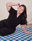 Ashley is sitting on a blue and white checkered rug wearing Petite Short Sleeve Jumpsuit in Basic Black