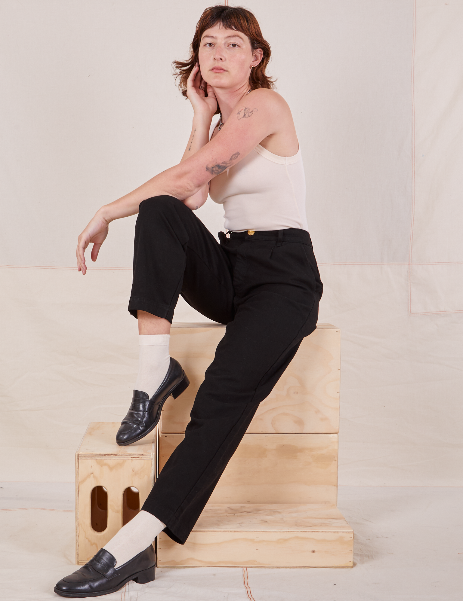 Alex is sitting on a stack of wooden crates wearing Denim Trouser Jeans in Black and Tank Top in vintage tee off-white