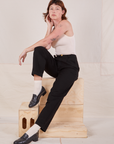 Alex is sitting on a stack of wooden crates wearing Denim Trouser Jeans in Black and Tank Top in vintage tee off-white