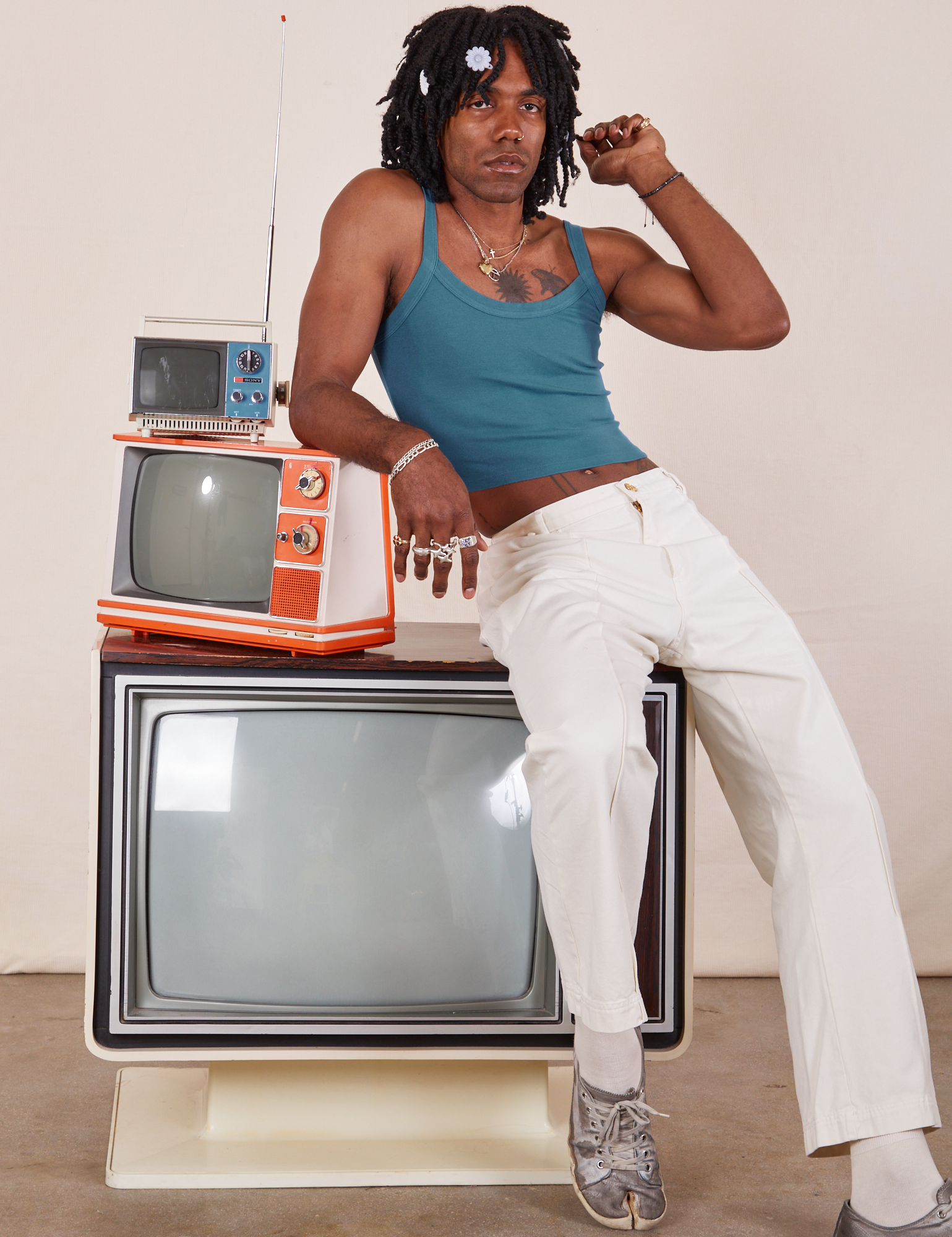 Three vintage tvs are stacked on top of each other. Jerrod is sitting on the large tv with his right elbow on a smaller orange and white tv. They are wearing Cropped Cami in Marine Blue and vintage tee off-white Western Pants.