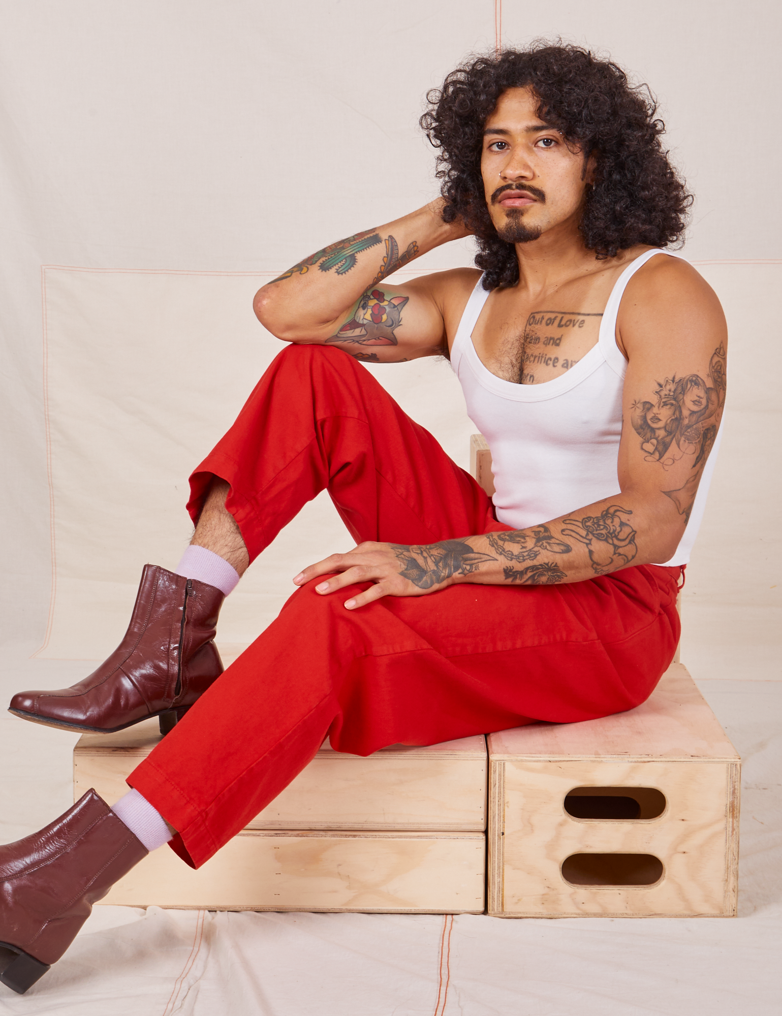 Jesse is sitting on a wooden crate. They are wearing Heavyweight Trousers in Mustang Red and Cropped Cami in vintage tee off-white
