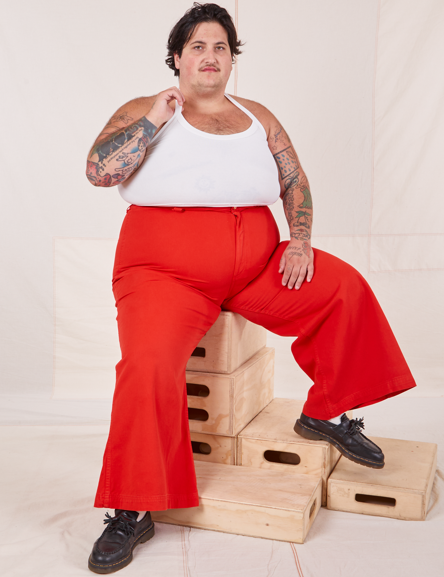 Sam is sitting on a stack of wooden crates. They are wearing Bell Bottoms in Mustang Red and Halter Top in vintage tee off-white