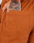 Back pocket close up of Short Sleeve Jumpsuit in Burnt Terracotta. Jesse has their hand in the pocket.