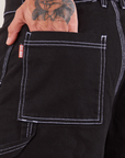 Carpenter Shorts in Basic Black back pocket close up. Jesse has their hand in the pocket.