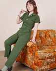 Hana is sitting on the arm of a sofa wearing Petite Short Sleeve Jumpsuit in Dark Emerald Green and holding the collar