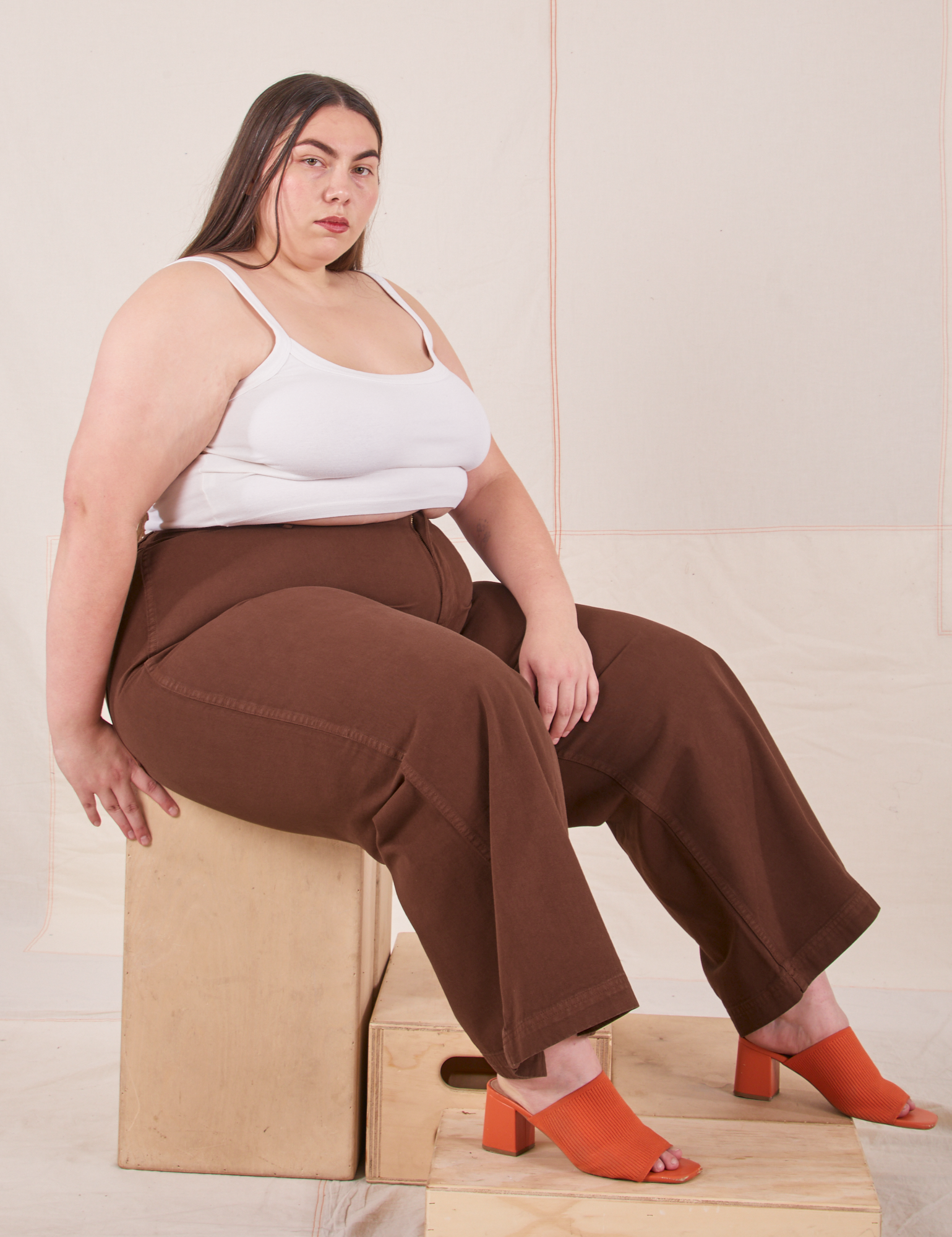Marielena is sitting on a wooden crate wearing Bell Bottoms in Fudgesicle Brown and Cropped Cami in vintage tee off-white