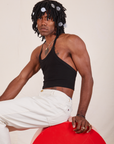 Jerrod is sitting on a red circular platform with his body facing the left side. They are wearing Halter Top in Basic Black and vintage off-white Western Pants