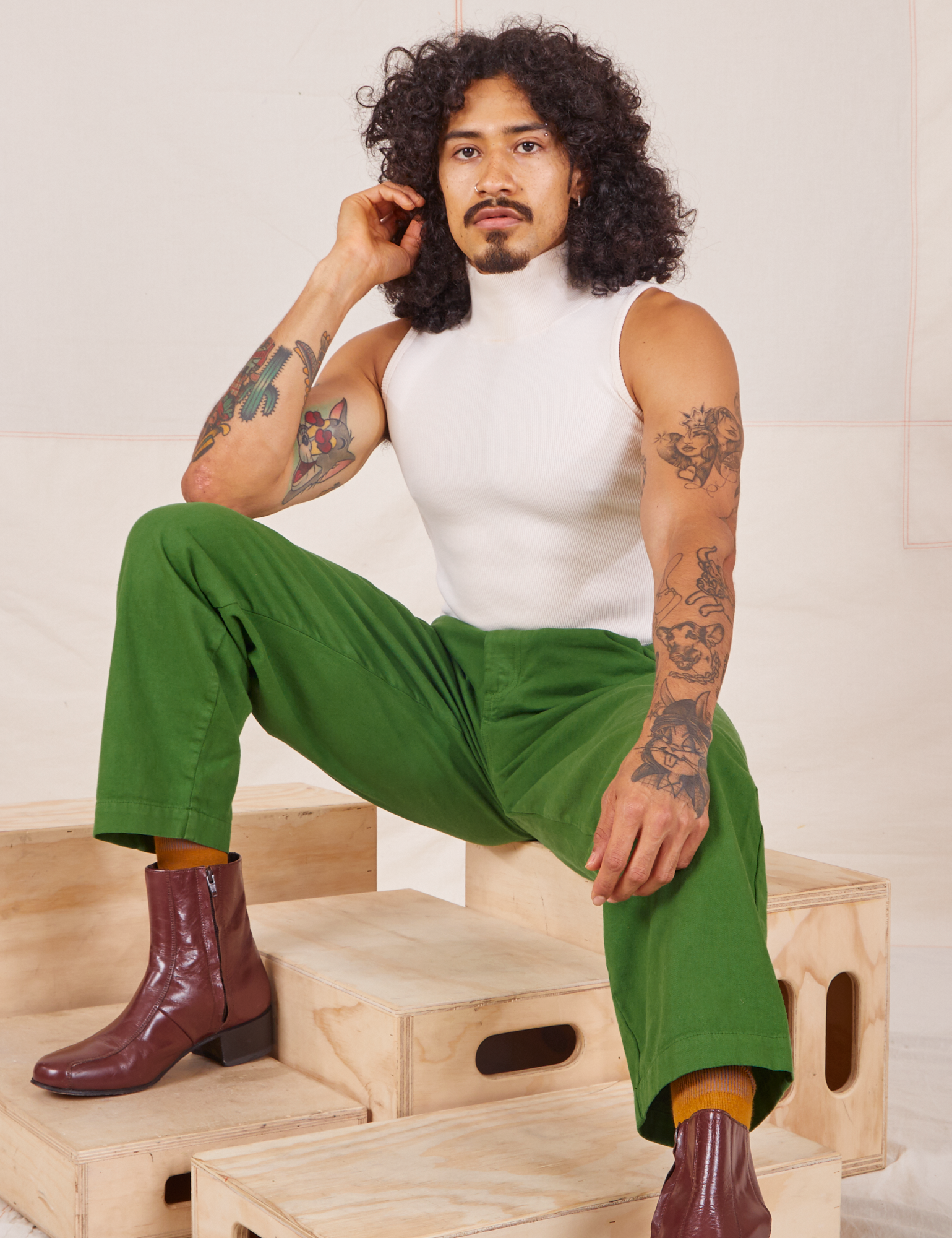 Jesse is sitting on a wooden crate wearing Heavyweight Trousers in Lawn Green and Sleeveless Turtleneck in vintage tee off-white