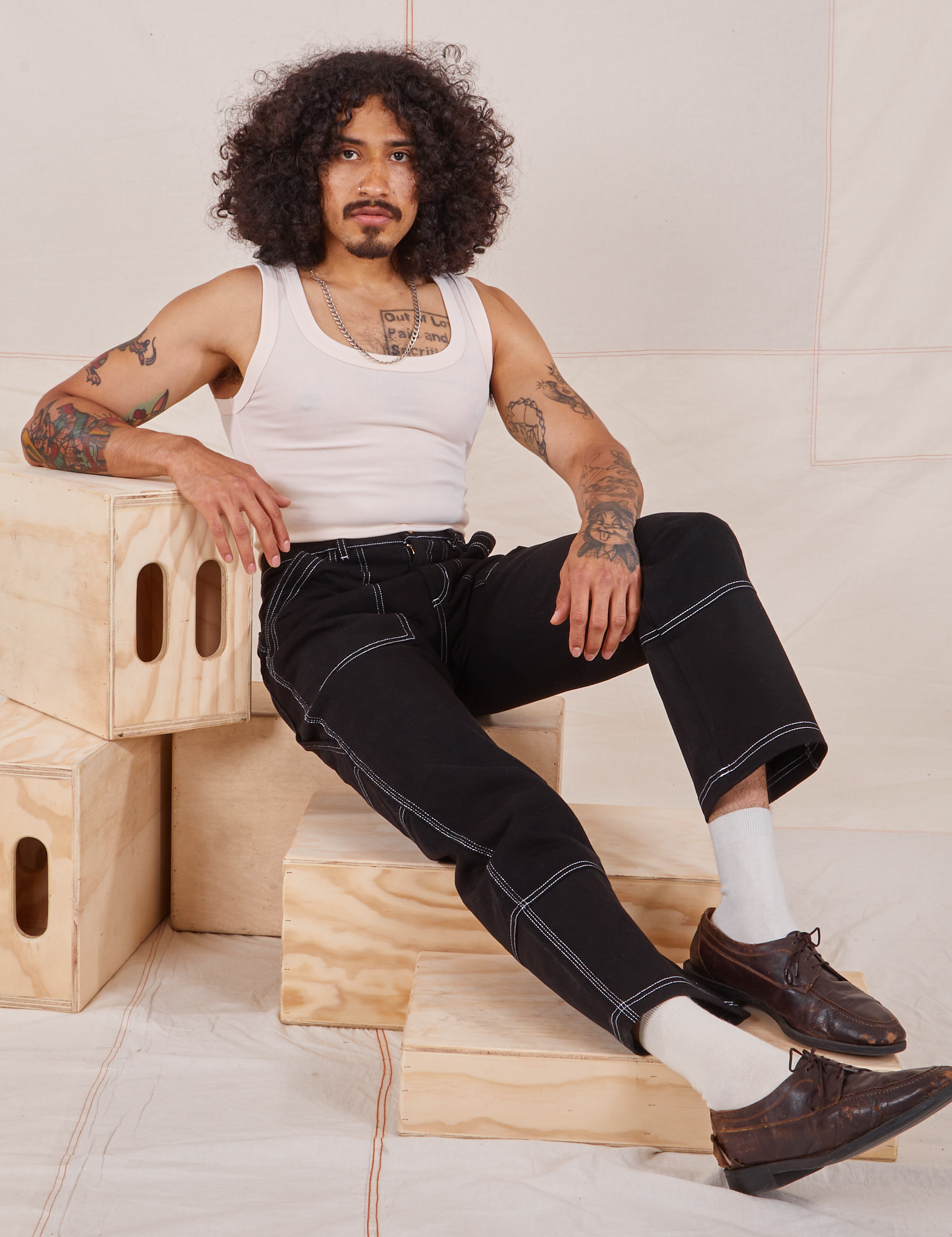 Jesse is sitting on a wooden crate. They are wearing Carpenter Jeans in Black and Tank Top in vintage tee off-white