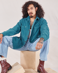 Jesse is wearing Corduroy Overshirt in Marine Blue and light wash Denim Trouser Jeans sitting on a wooden crate