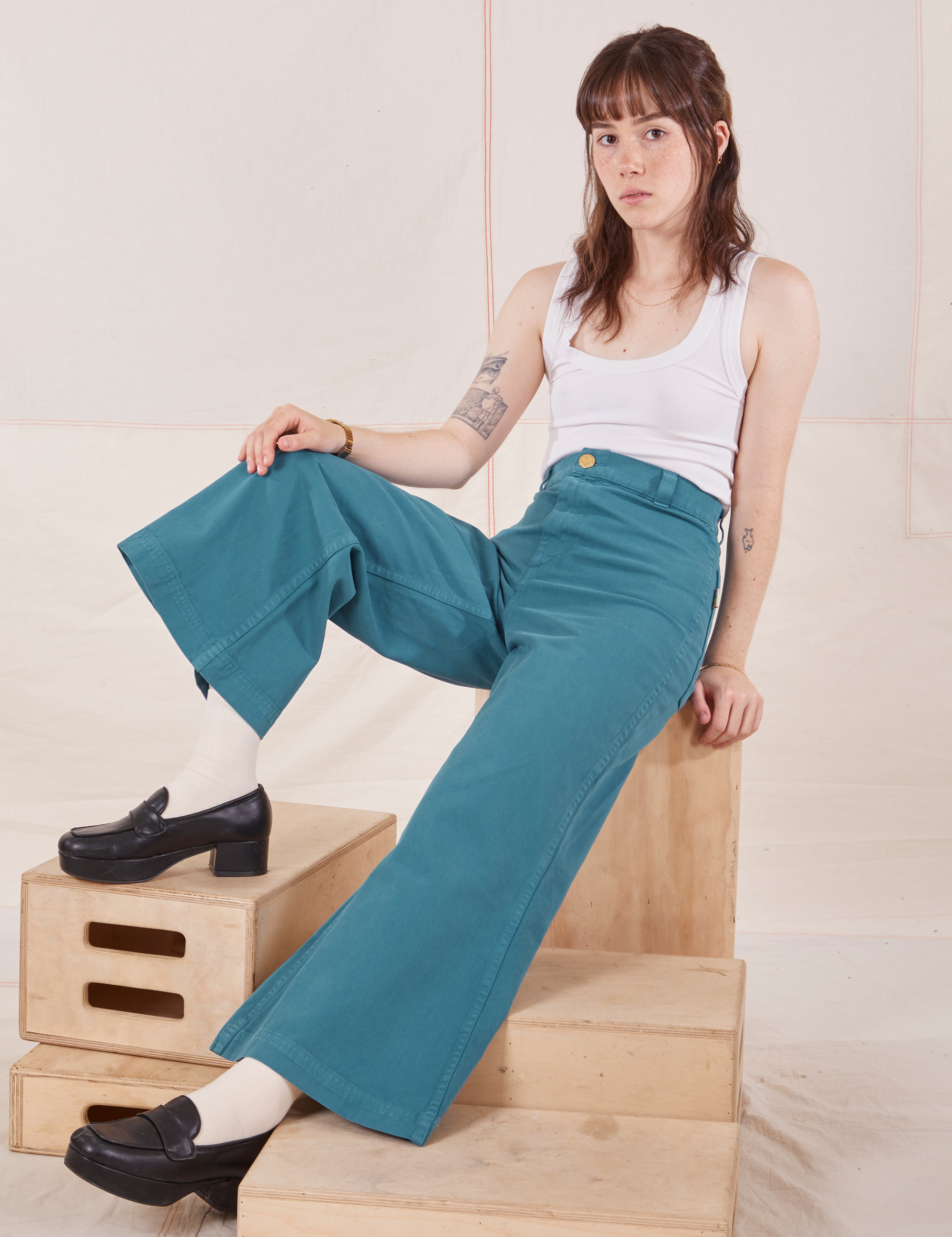 Hana is sitting on a wooden crate wearing Petite Bell Bottoms in Marine Blue and Cropped Tank Top in vintage tee off-white