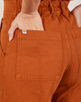Back pocket close up of Short Sleeve Jumpsuit in Burnt Terracotta. Tiara has her hand in the pocket.