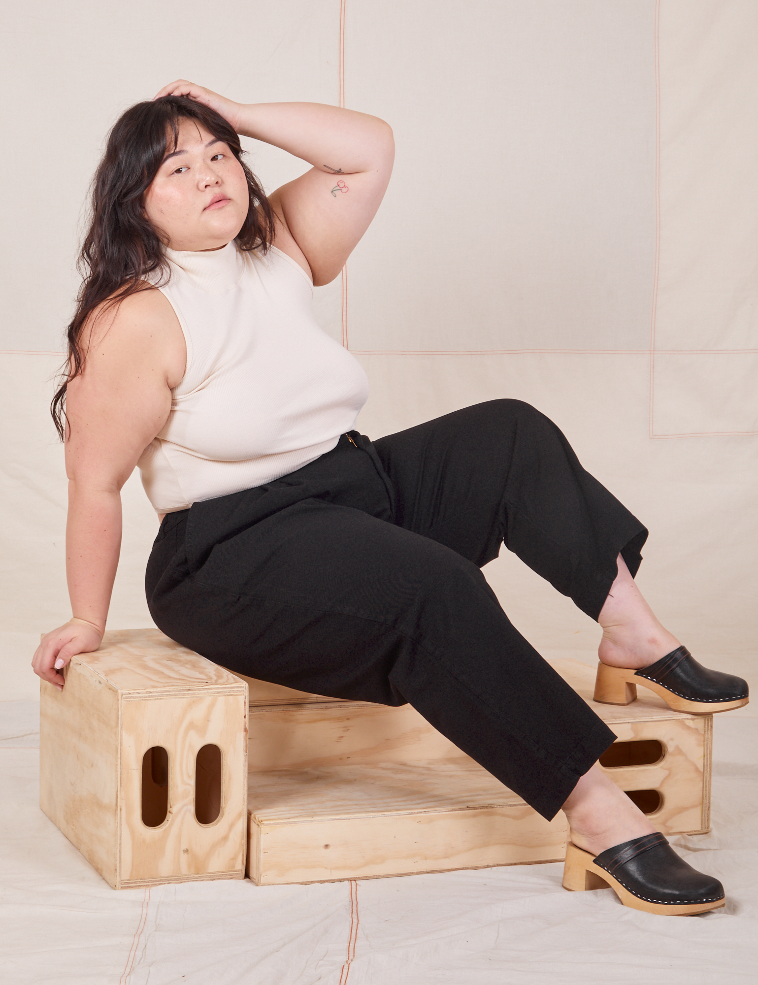 Ashley is wearing Heavyweight Trousers in Basic Black and Sleeveless Turtleneck in vintage tee off-white. She is sitting on a wooden crate.