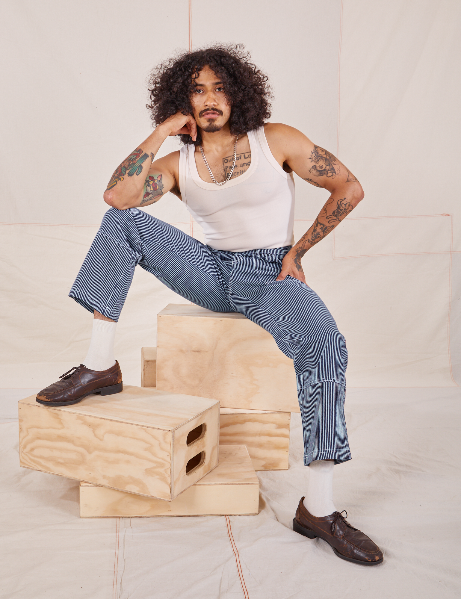 Jesse is sitting on a stack of wooden crates. They are wearing Carpenter Jeans in Railroad Stripes and Tank Top in vintage tee off-white