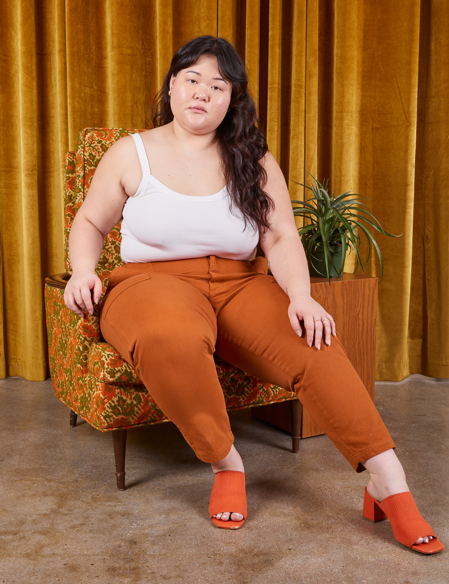 Ashley is sitting on a upholstered chair wearing Petite Pencil Pants in Burnt Terracotta and Cropped Cami in vintage tee off-white