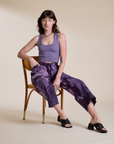 Alex is sitting in a chair and wearing Magic Waters Drop Pocket Pants in Dark Lavender paired with faded grape Cropped Tank