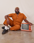 Elijah is wearing Short Sleeve Jumpsuit in Burnt Terracotta and sitting next to a small vintage tv.