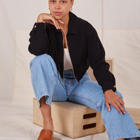 Gabi is sitting on a wooden crate wearing Ricky Jacket in Basic Black