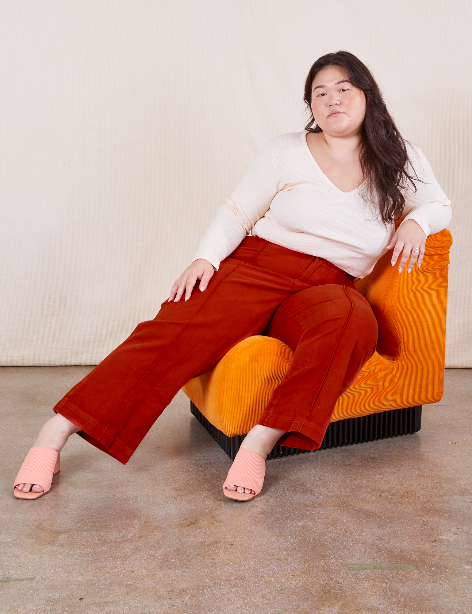 Ashley is sitting on an orange upholstered chair. She is wearing Western Pants in Paprika paired with a Long Sleeve V-Neck Tee in vintage tee off-white