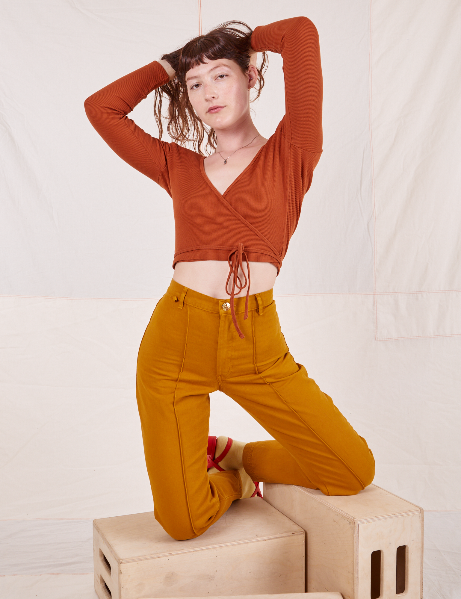 Alex is wearing Wrap Top in Burnt Terracotta and spicy mustard Western Pants and is kneeling on wooden crates