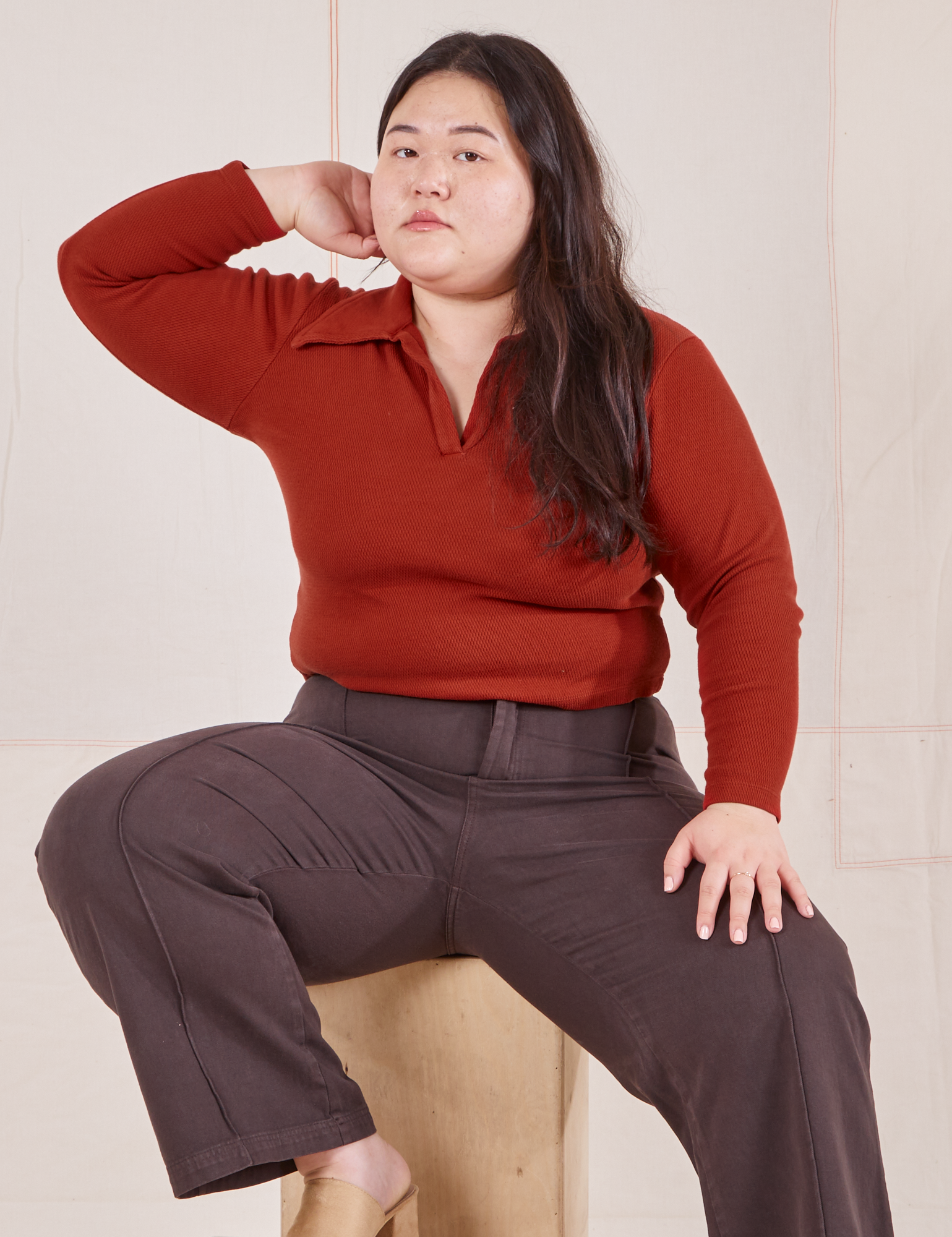 Ashley is sitting on a wooden crate wearing Long Sleeve Fisherman Polo in Paprika and espresso brown Western Pants