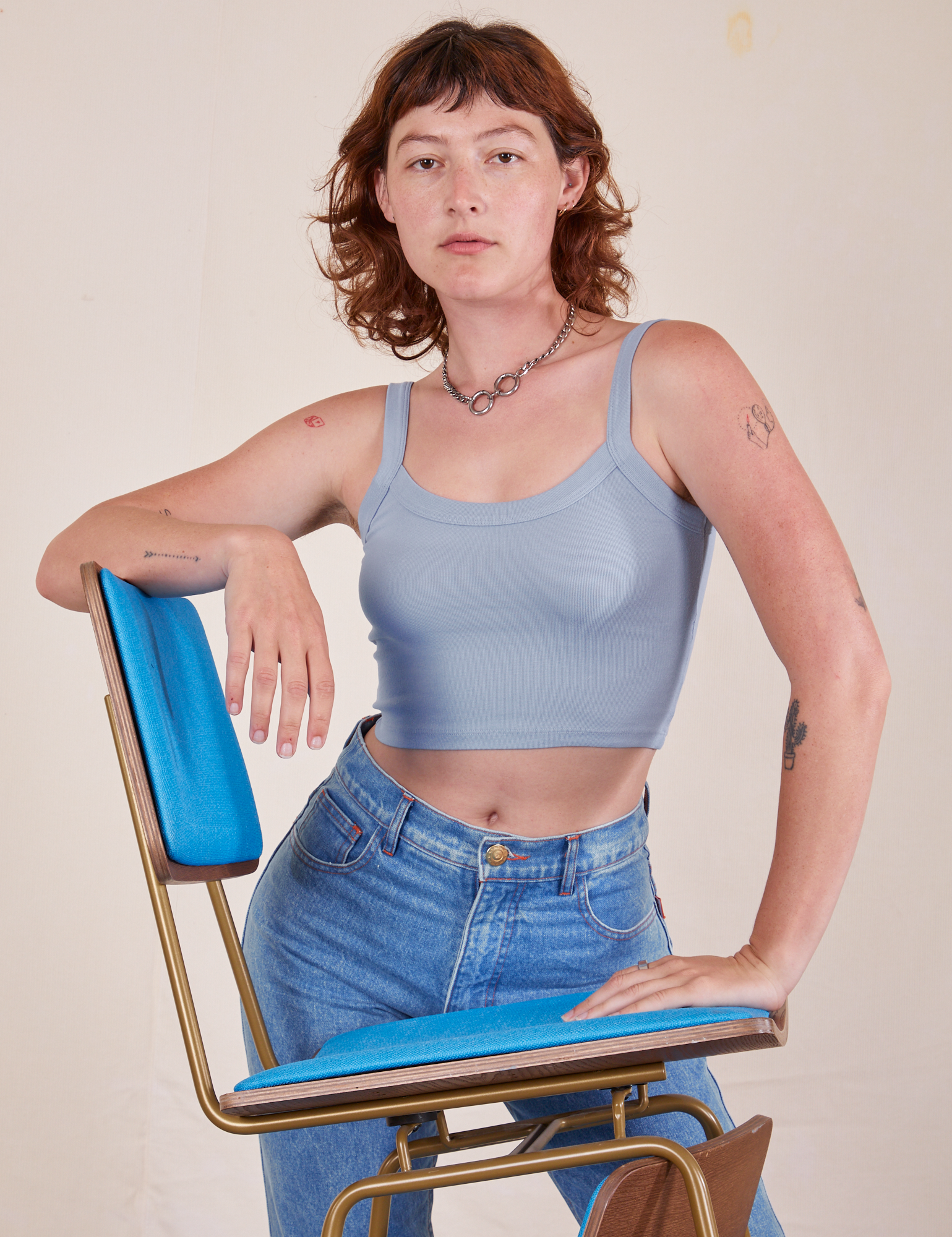 Alex is 5'8" and wearing P Cropped Cami in Periwinkle. There are two blue and brass vintage chairs stacked on top of each other. Alex has her elbow on the top back of one chair and the other hand on the seat.
