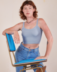 Alex is 5'8" and wearing P Cropped Cami in Periwinkle. There are two blue and brass vintage chairs stacked on top of each other. Alex has her elbow on the top back of one chair and the other hand on the seat.