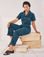 Mika is wearing Heritage Short Sleeve Jumpsuit in Lagoon and sitting on a stack of wooden cartons