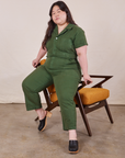 Ashley is sitting on the arm of a chair wearing Petite Short Sleeve Jumpsuit in Dark Emerald Green