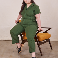 Ashley is sitting on the arm of a chair wearing Petite Short Sleeve Jumpsuit in Dark Emerald Green