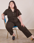 Ashley is sitting in a blue and brass chair wearing Petite Short Sleeve Jumpsuit in Espresso Brown 