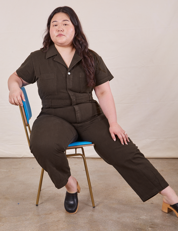 Ashley is sitting in a blue and brass chair wearing Petite Short Sleeve Jumpsuit in Espresso Brown 