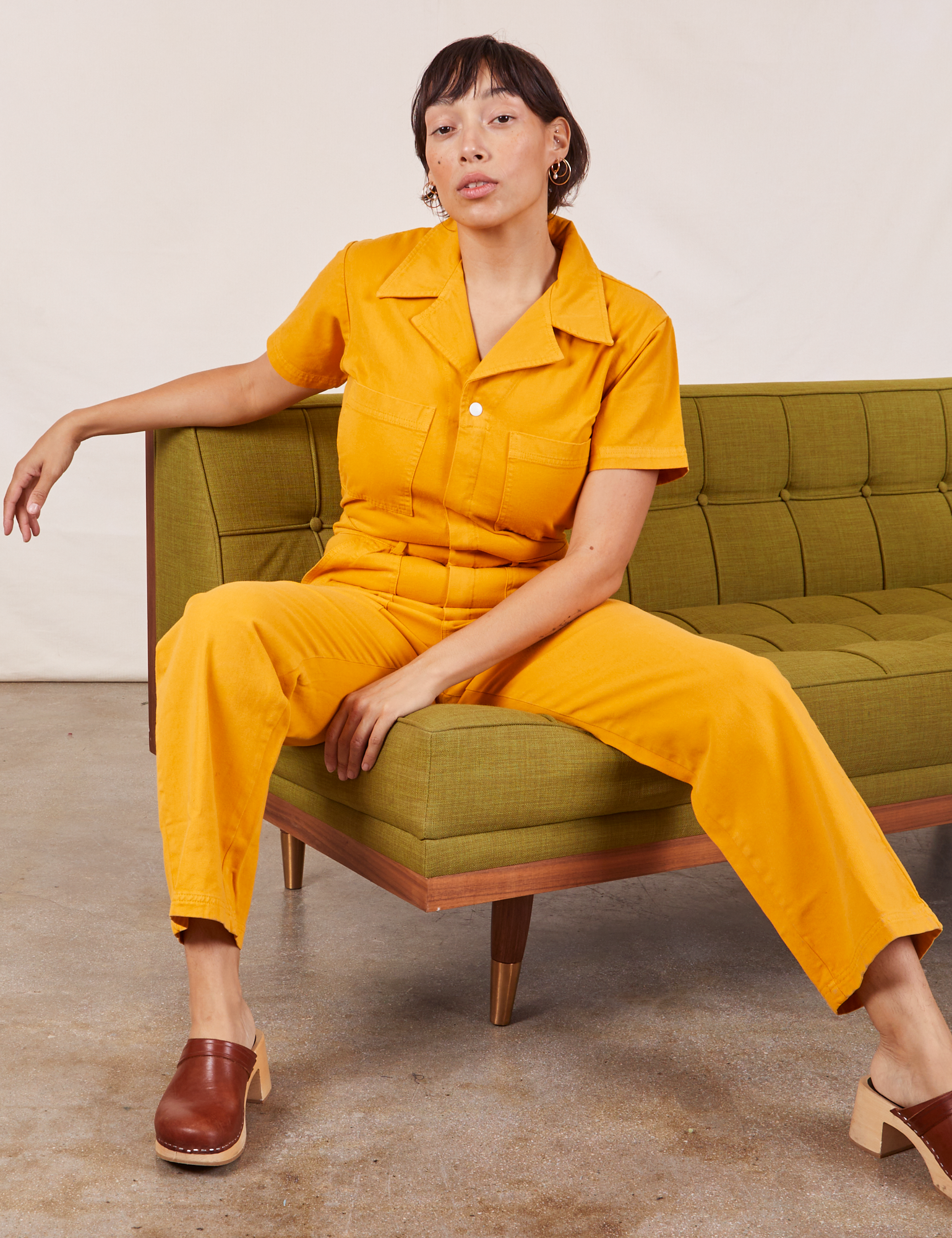Tiara is sitting on a green couch wearing Short Sleeve Jumpsuit in Mustard Yellow