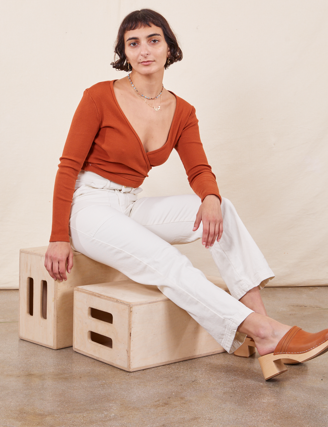 Soraya is sitting on a wooden crate wearing Work Pants in Vintage Off-White and burnt terracotta Wrap Top