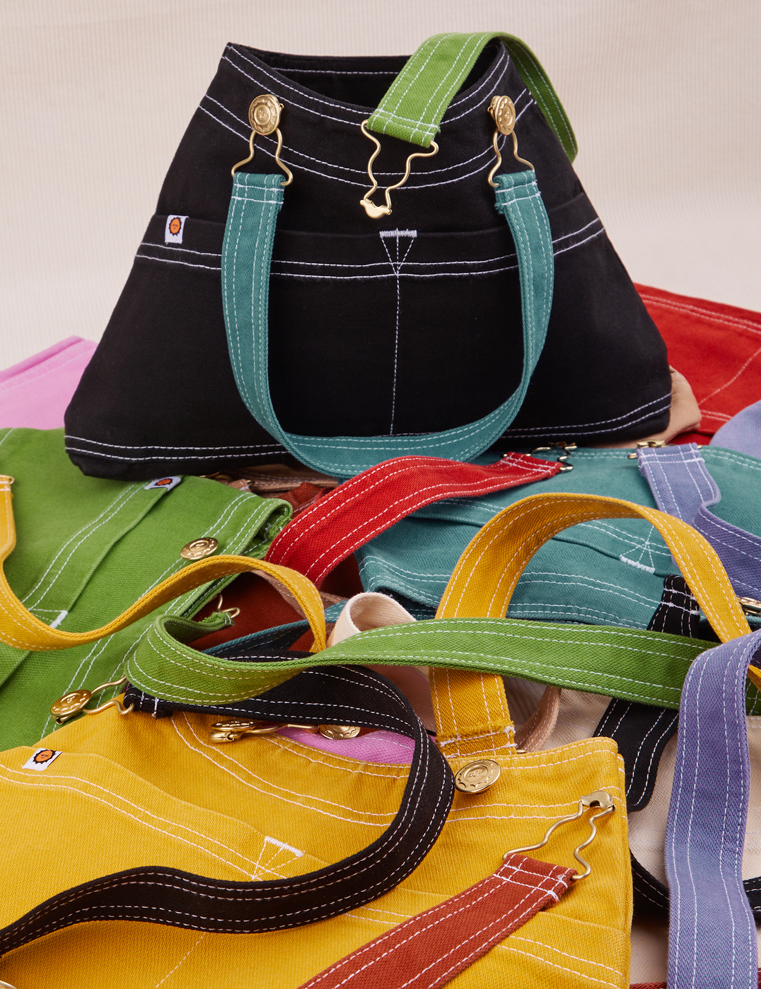 Overall Handbag in an array of colors. Black standing up with marine blue strap. All other bags laid flat underneath and around black bag.