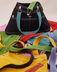 Overall Handbag in an array of colors. Black standing up with marine blue strap. All other bags laid flat underneath and around black bag.