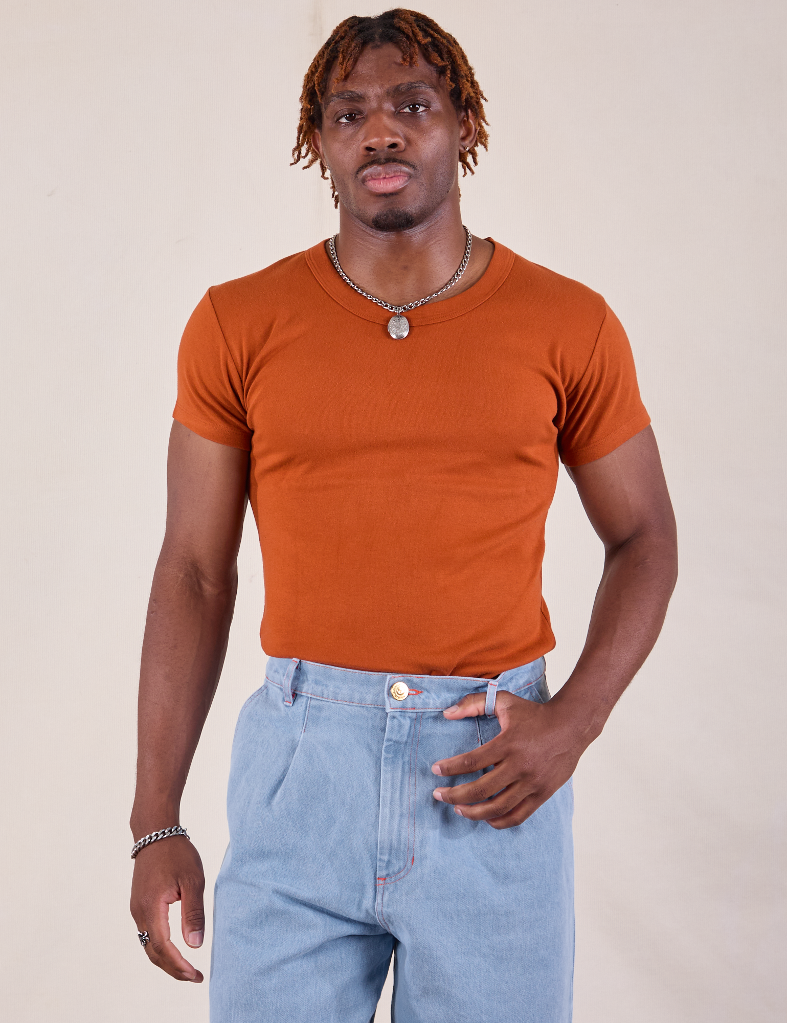 Isaac is wearing Baby Tee in Burnt Terracotta tucked into light wash Denim Trousers