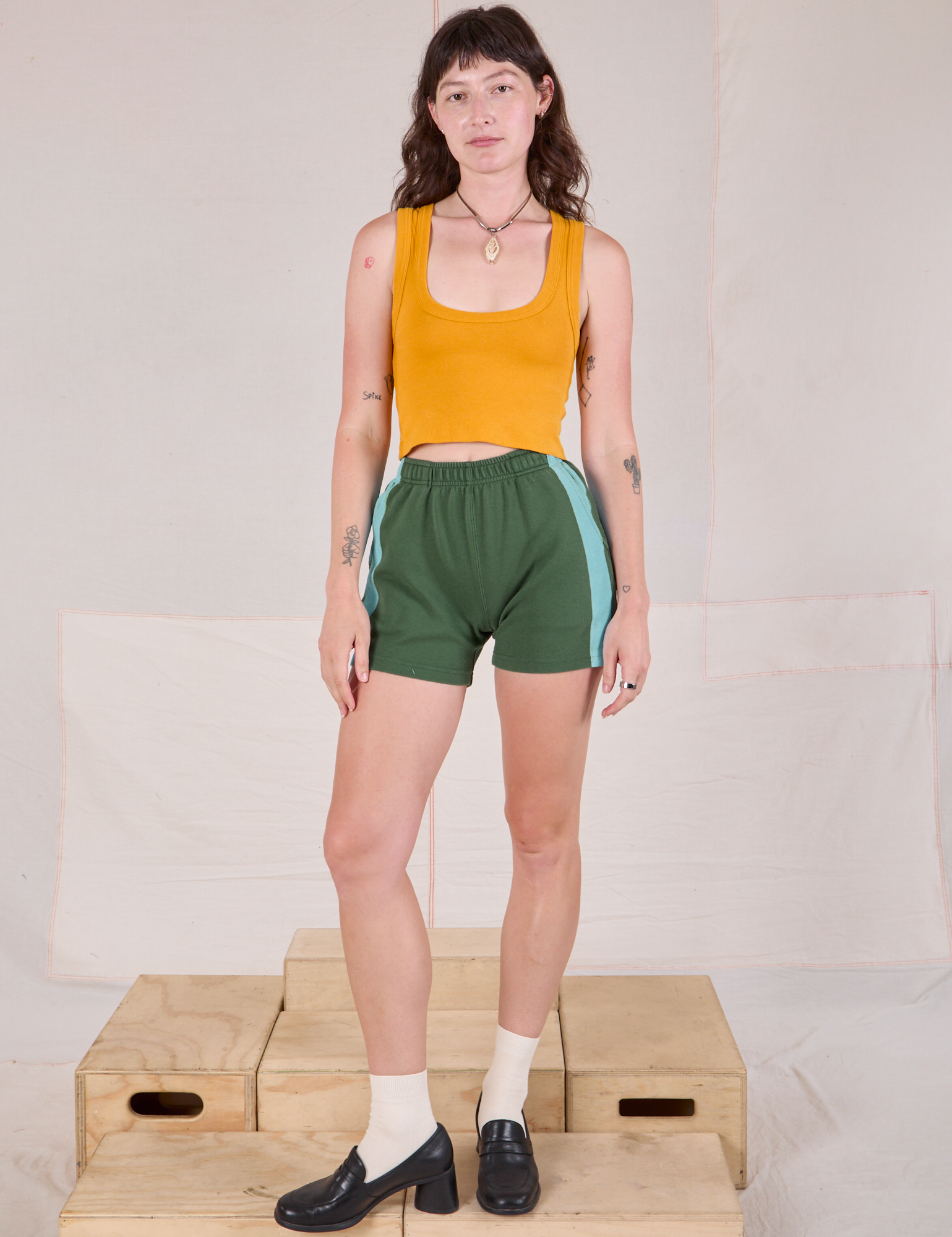Alex is 5'8" and wearing P Painted Stripe Easy Shorts in Dark Emerald Green paired with a golden yellow Cropped Tank