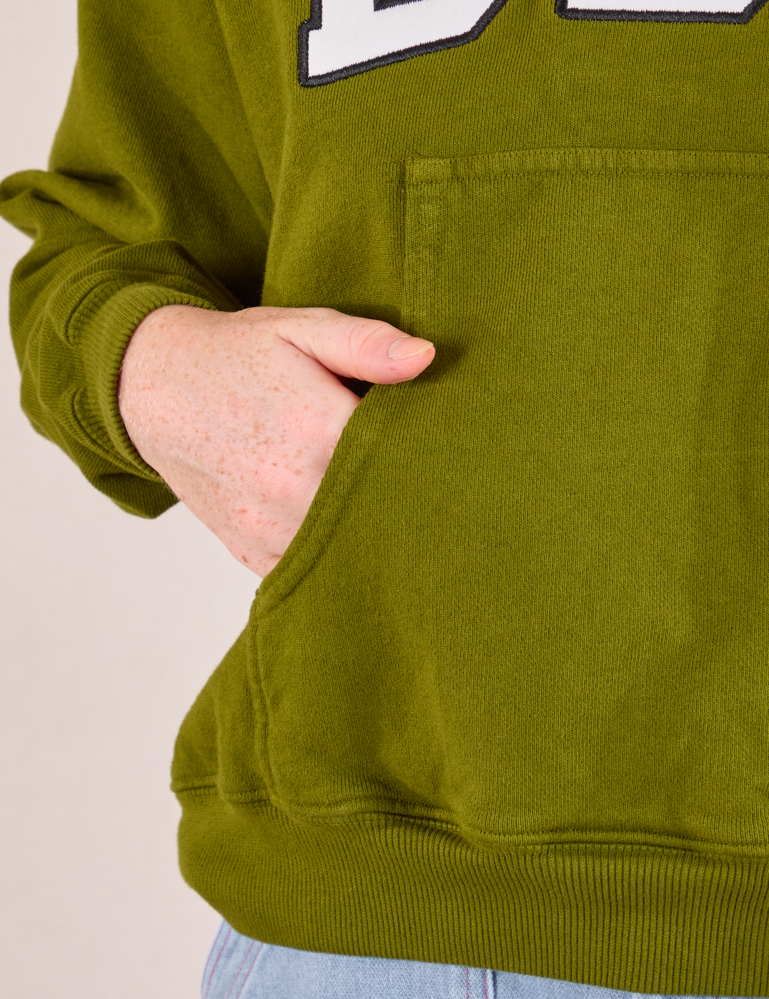 Collegiate Hoodie in Summer Olive kangaroo pocket close up. Margaret has her hand in the pocke.