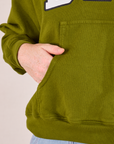 Collegiate Hoodie in Summer Olive kangaroo pocket close up. Margaret has her hand in the pocke.