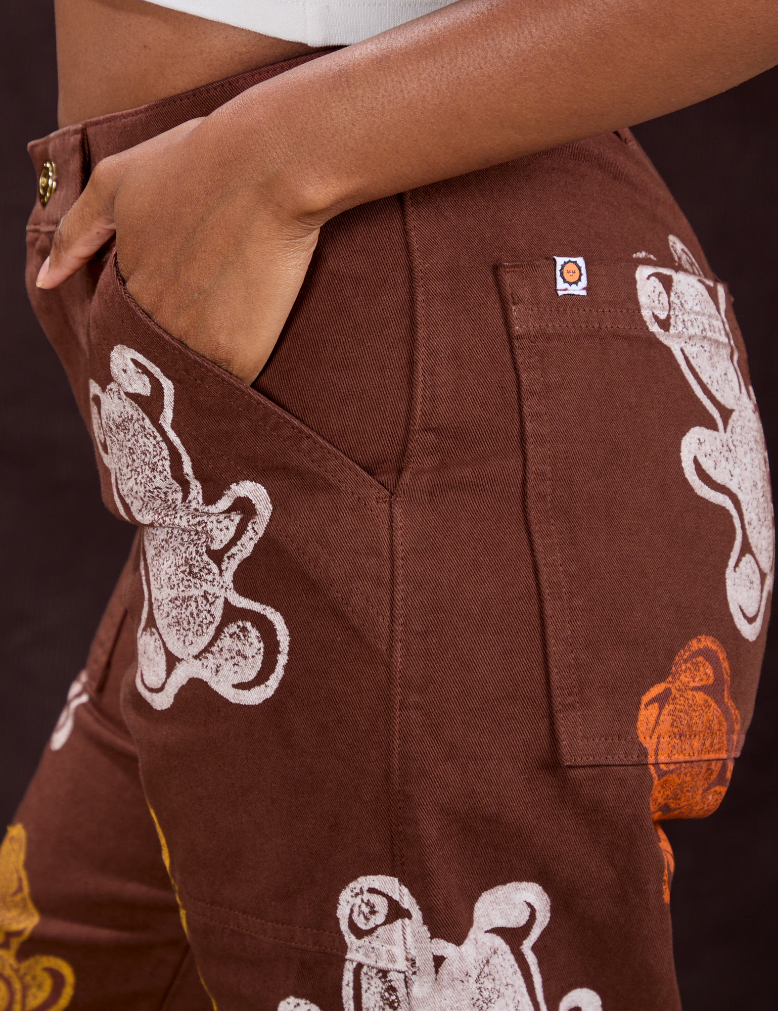 Close up of Kandia&#39;s hand in the front pocket of Gummy Bear Paintstamped Work Pants in Fudgesicle Brown