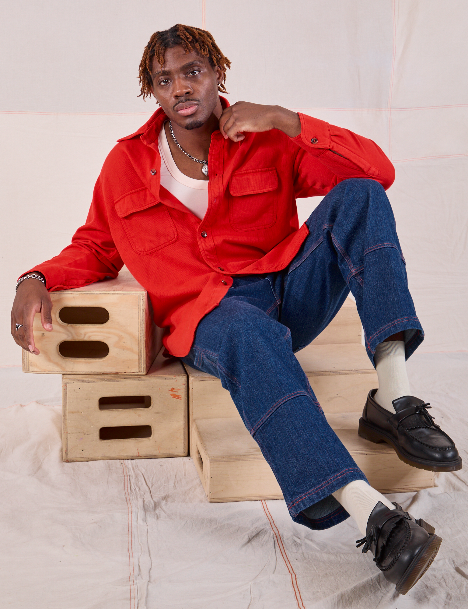 Flannel Overshirt in Mustang Red and dark wash Carpenter Jeans worn by Isaac sitting on a wooden box.