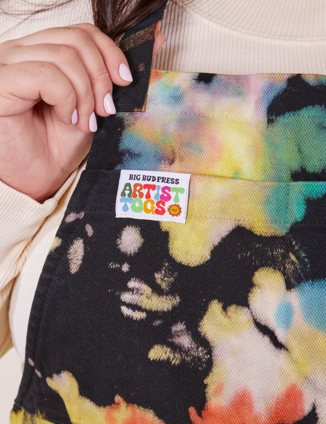 Front close up of Artist Togs Full Apron in Rainbow Magic Waters. Ashley is holding onto the neck strap. Label sewn on front of apron that reads "Big Bud Press" in black text and "Artist Togs" in rainbow text.