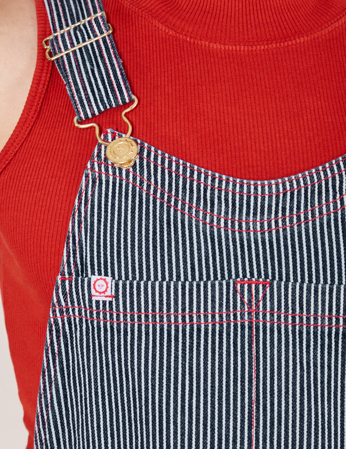 Front close up of Railroad Stripe Denim Original Overalls. Red contrast stitching and white and red sun baby tag.