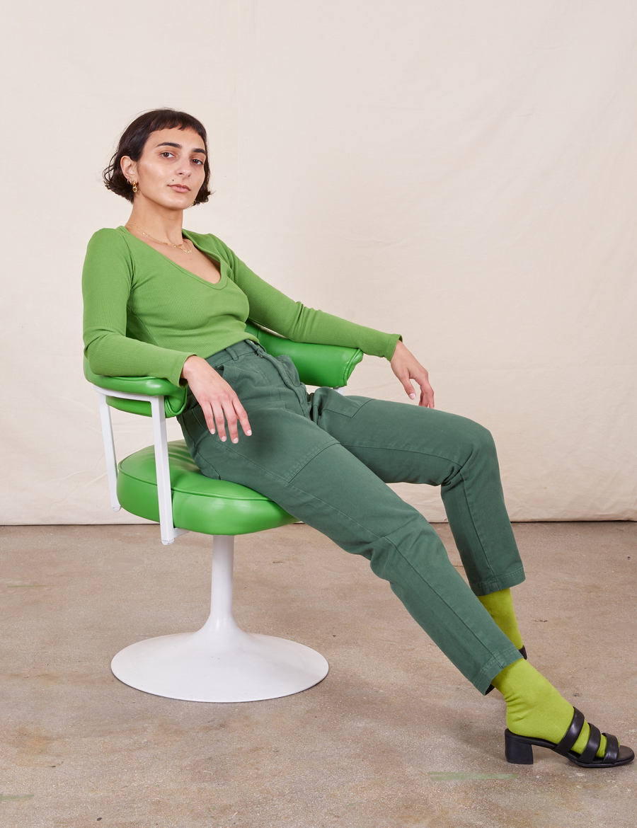 Soraya is sitting in a green chair with white frame. She is wearing Pencil Pants in Dark Emerald Green paired with a bright olive Long Sleeve V-Neck Tee