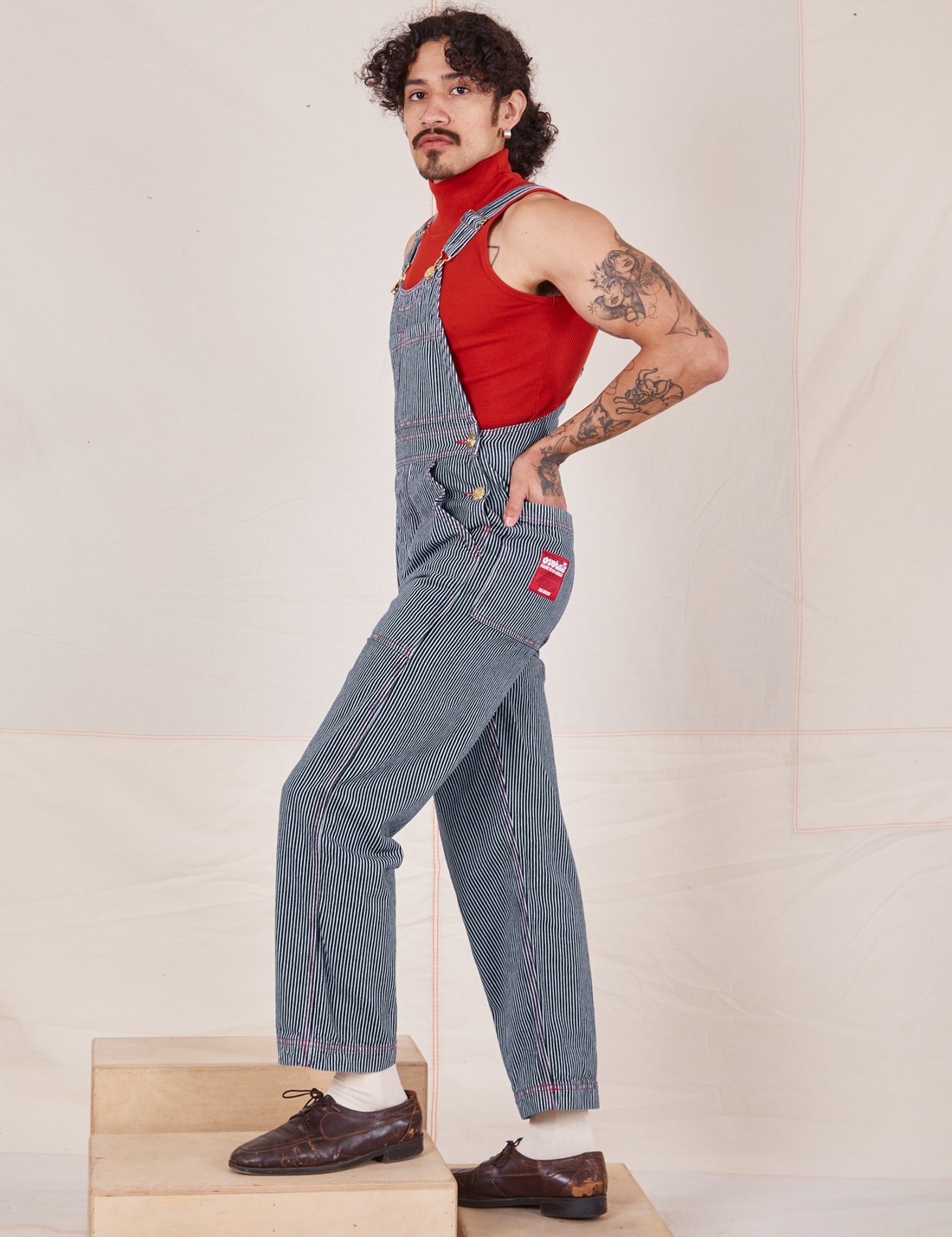 Side view of Railroad Stripe Denim Original Overalls and paprika Sleeveless Turtleneck worn by Jesse. They have their hand in the back pocket