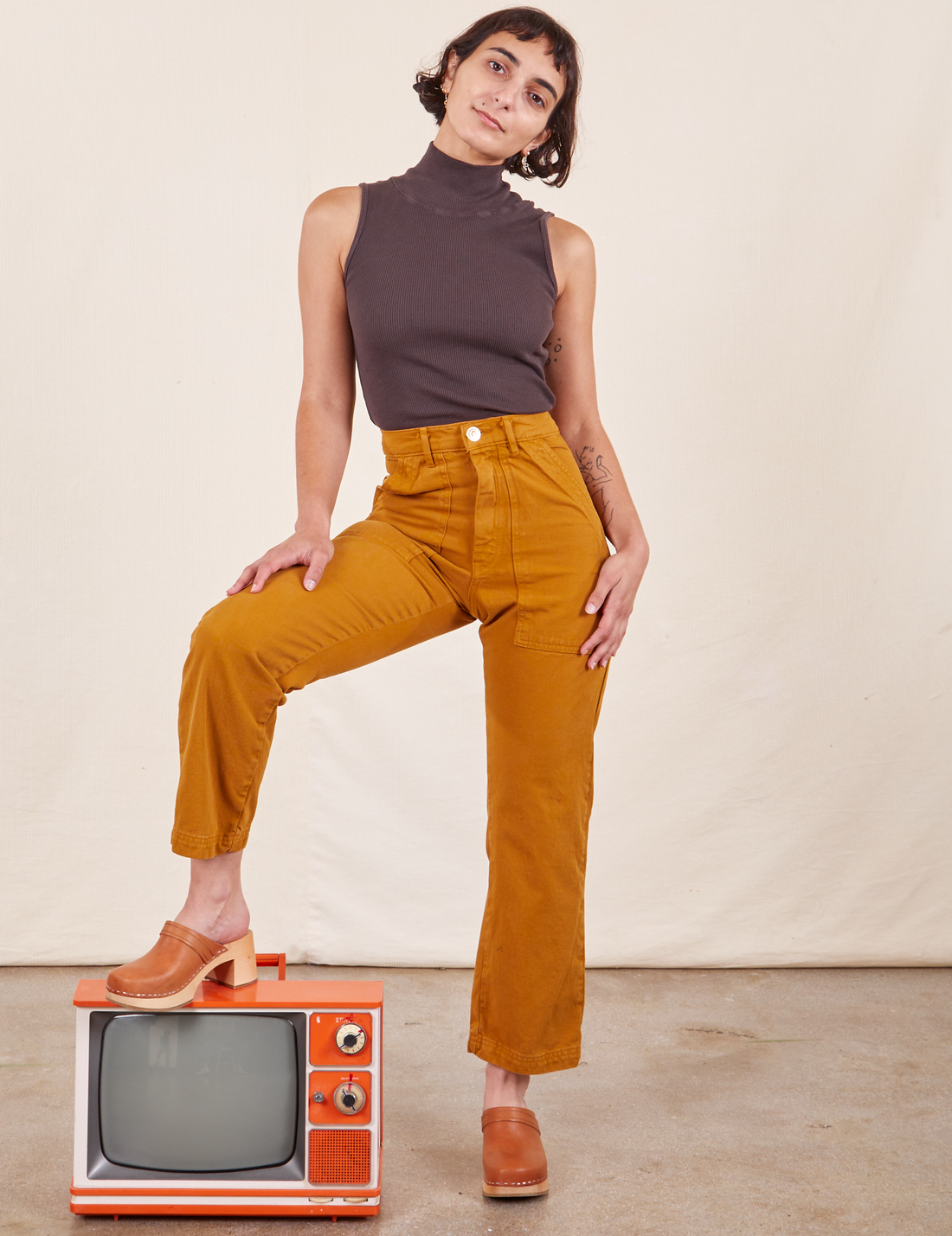 Work Pants in Spicy Mustard on Soraya wearing espresso brown Sleeveless Turtleneck. She has one foot on a small orange vintage tv with the other foot on the ground.