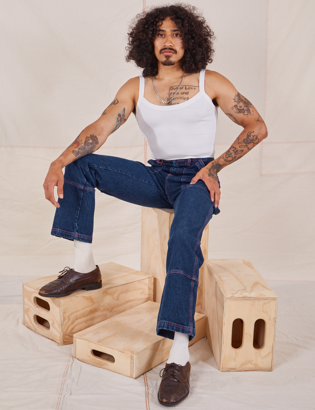 Jesse is sitting on a wooden crate. They are wearing Carpenter Jeans in Dark Wash and a vintage off-white Cami