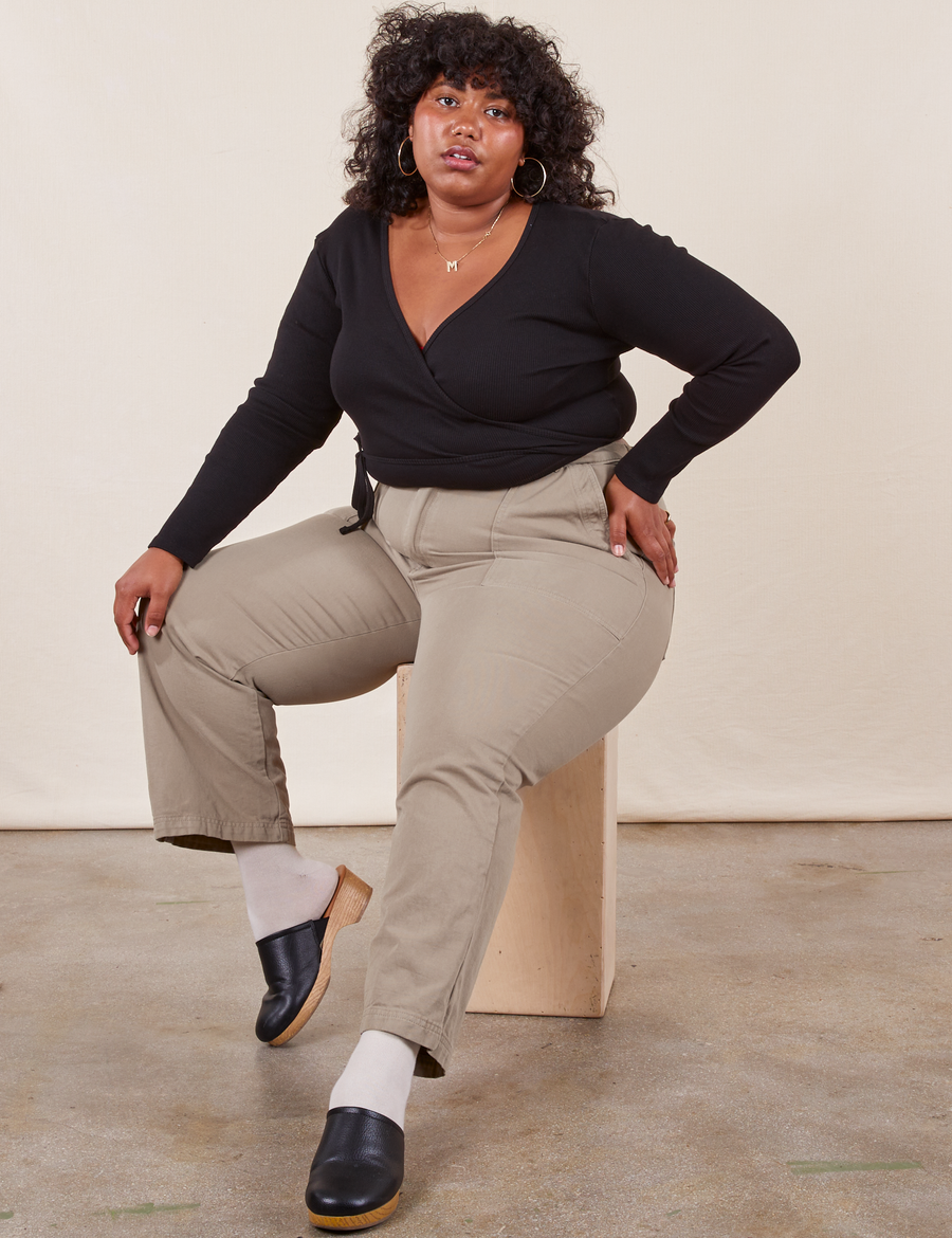 Morgan is sitting on a wooden crate wearing Work Pants in Khaki Grey and a black Wrap Top