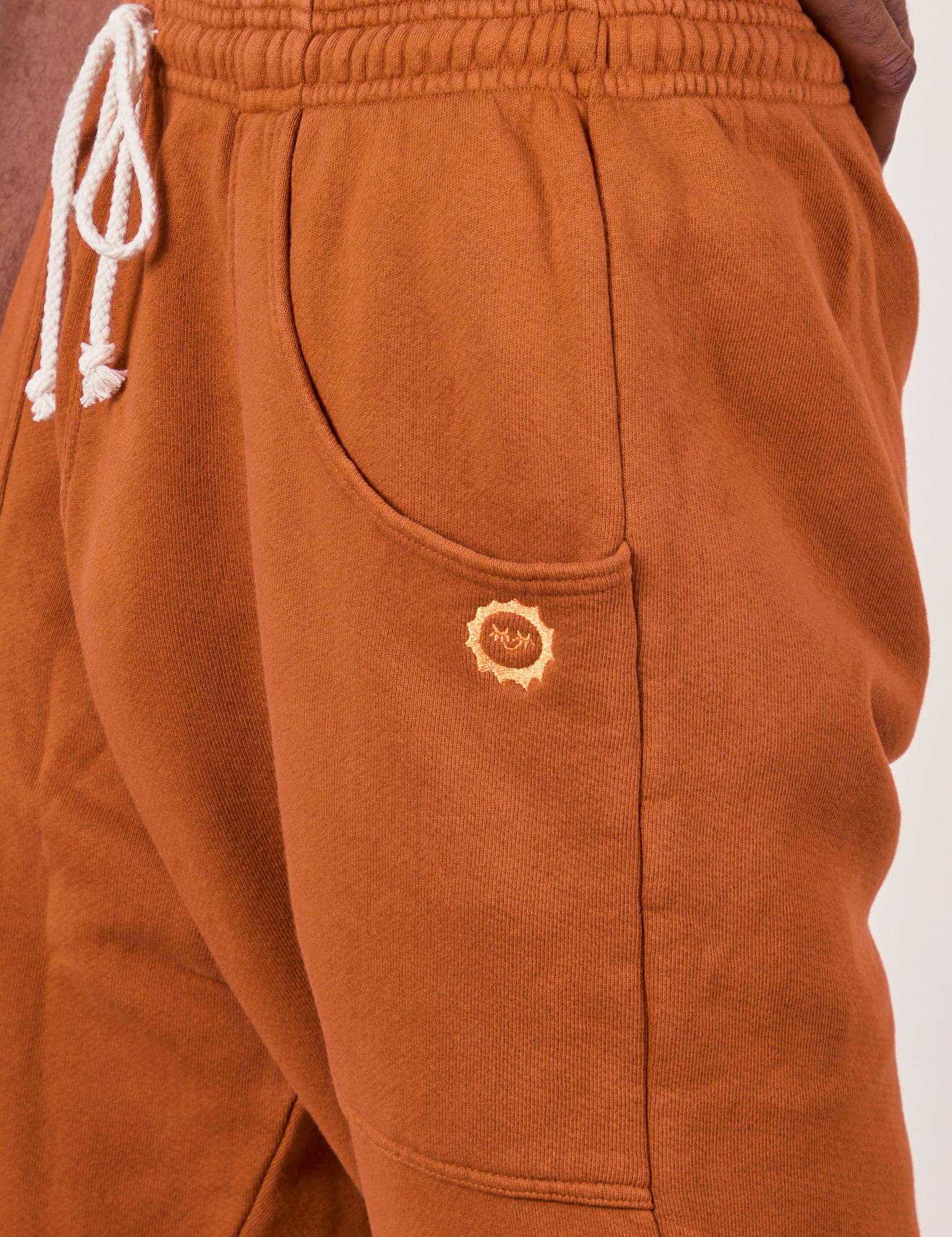 Side view close up of Cropped Rolled Cuff Sweatpants in Burnt Terracotta on Jerrod. Embroidered Sun Baby logo on pocket.